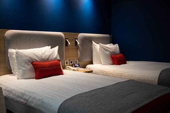 A hotel room with two beds, each with white bedding and red pillows. There is a wooden headboard, a bedside table with a lamp, and a wall-mounted phone. - Holiday Inn Express London Stansted Airport by IHG (Bishop's Stortford)