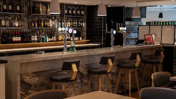 Uma área de bar com bancada de mármore, diversas garrafas de bebidas e banquetas.