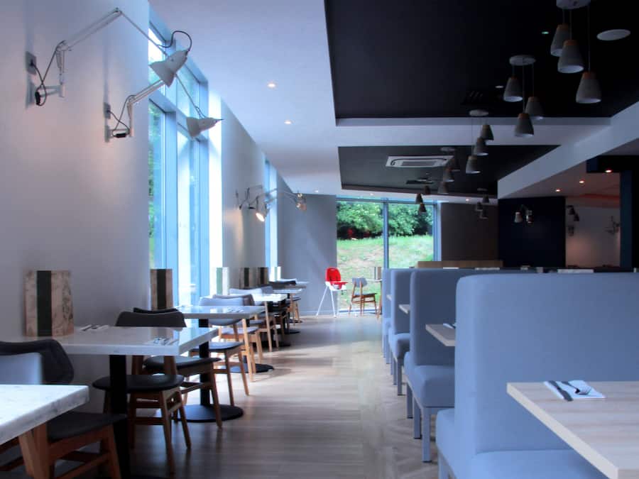 Interior moderno de restaurante con mesas altas, cabinas, ventanas amplias e iluminación colgante.