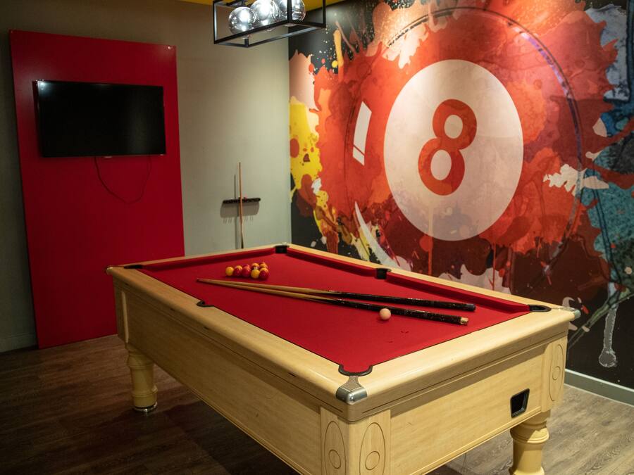 A pool table with balls and cues, a red wall, a television mounted on a red panel, and a colorful abstract mural with a large number '8'.