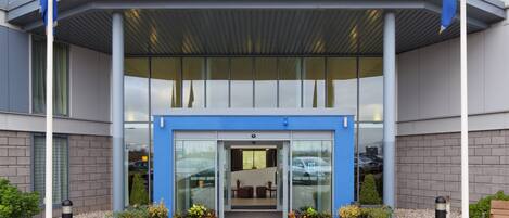 Fachada moderna de un edificio con toldo azul, puertas de vidrio y plantas en macetas.