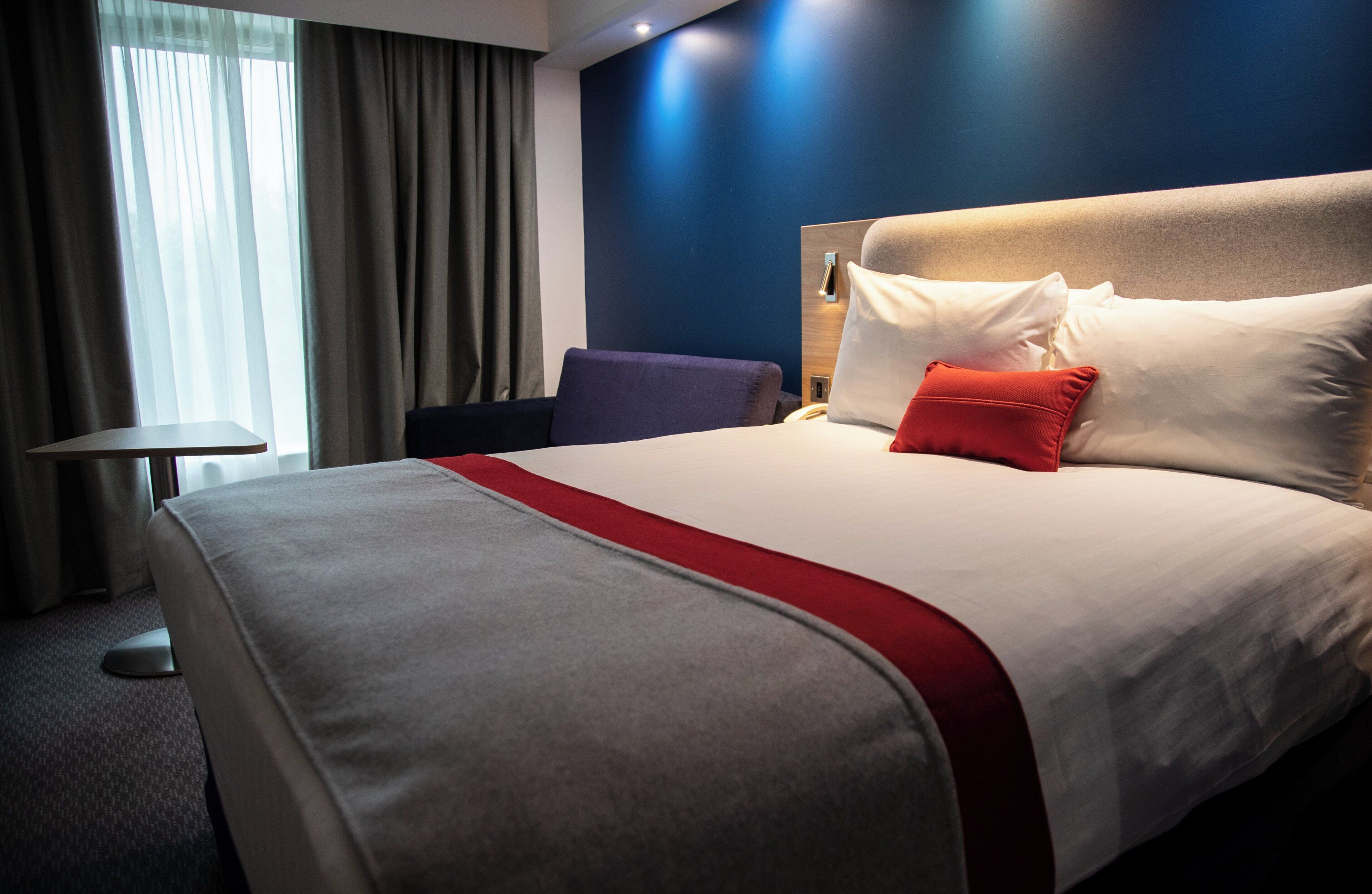 Ein Hotelzimmer mit einem großen Bett, einem Nachttisch, einem Sessel und einem Fenster mit Vorhängen.