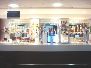 Een bar met verschillende drankdispensers, een bord waarop staat dat bestellingen alleen via de bar kunnen, en een verscheidenheid aan flessen en glazen.