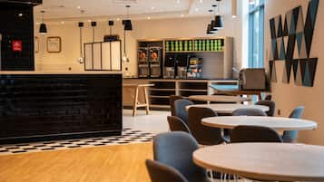 Interior de una cafetería moderna con mostrador de ladrillo negro, zona de estar con mesas y sillas redondas, una máquina de café y una pared con arte geométrico.