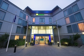 A Holiday Inn Express building with a modern facade, large windows, and a well-lit entrance. - Holiday Inn Express London Stansted Airport by IHG (Bishop's Stortford)