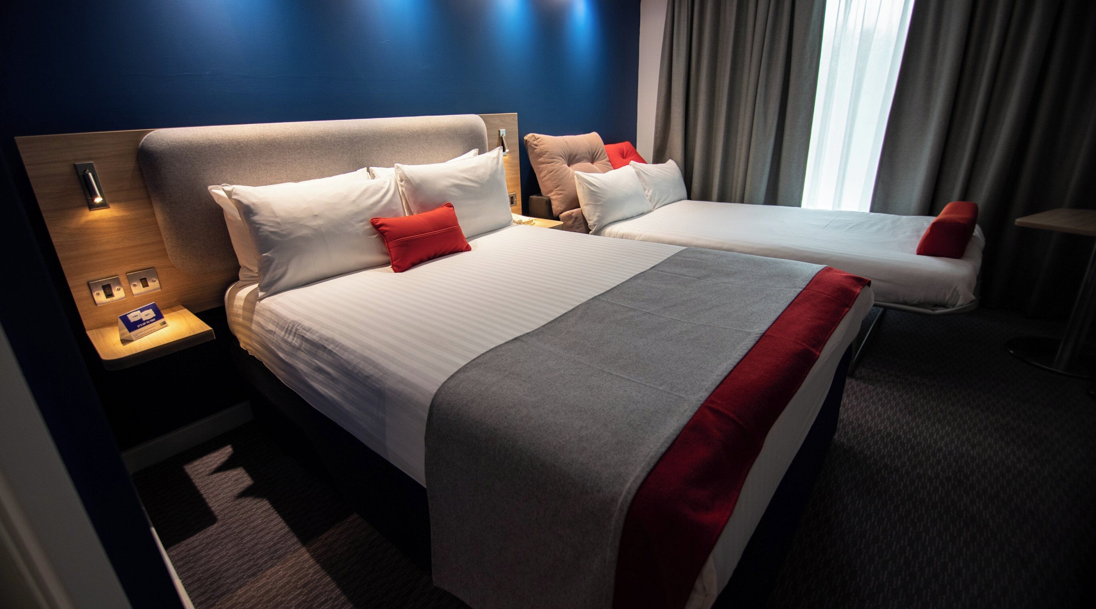 A hotel room with two beds, a nightstand, a lamp, and a window with curtains.