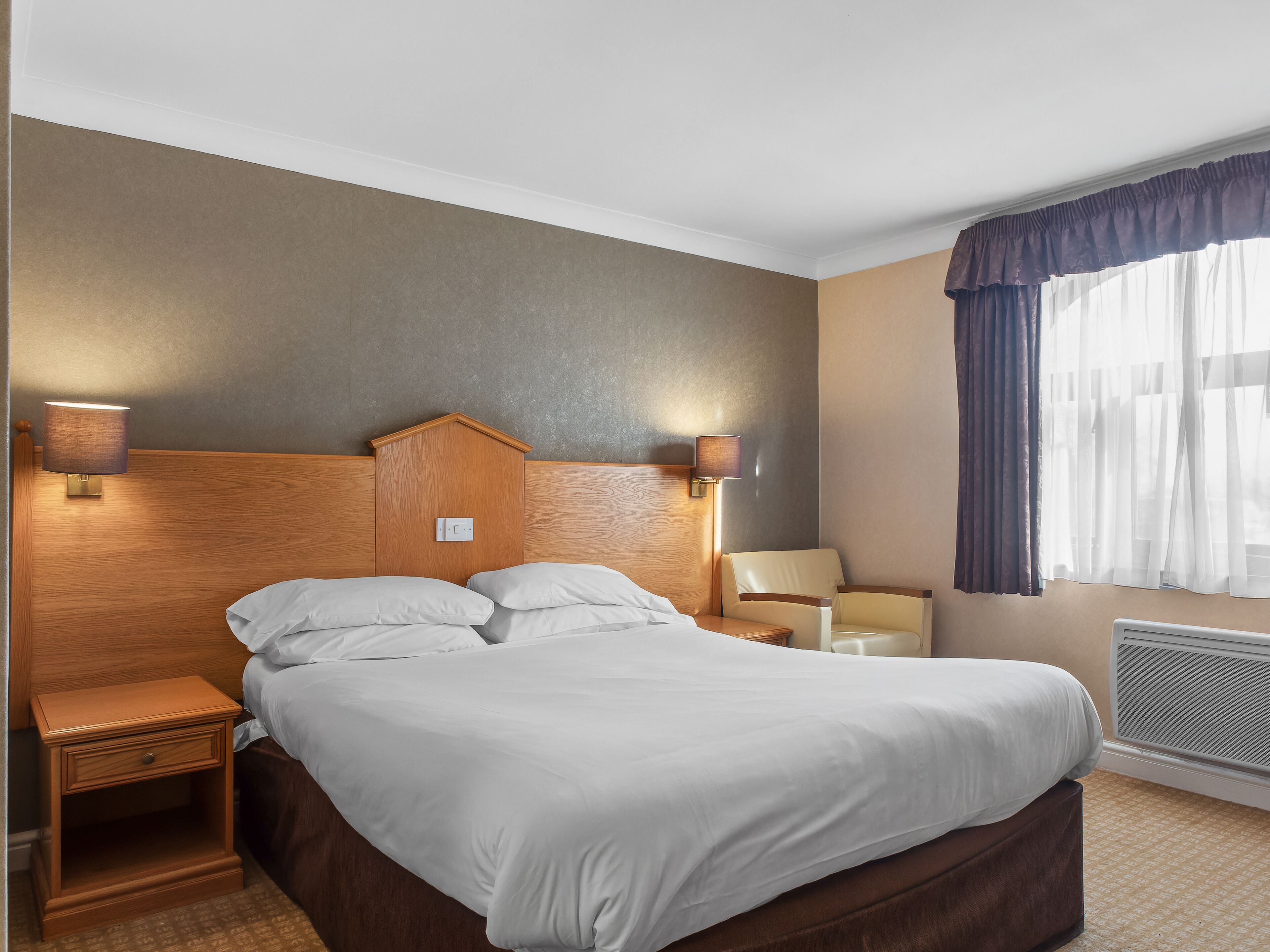 Una habitación de hotel con cabecera de madera, cama con sábanas blancas, dos lámparas de noche, una mesita de noche, una silla y una ventana con cortinas.
