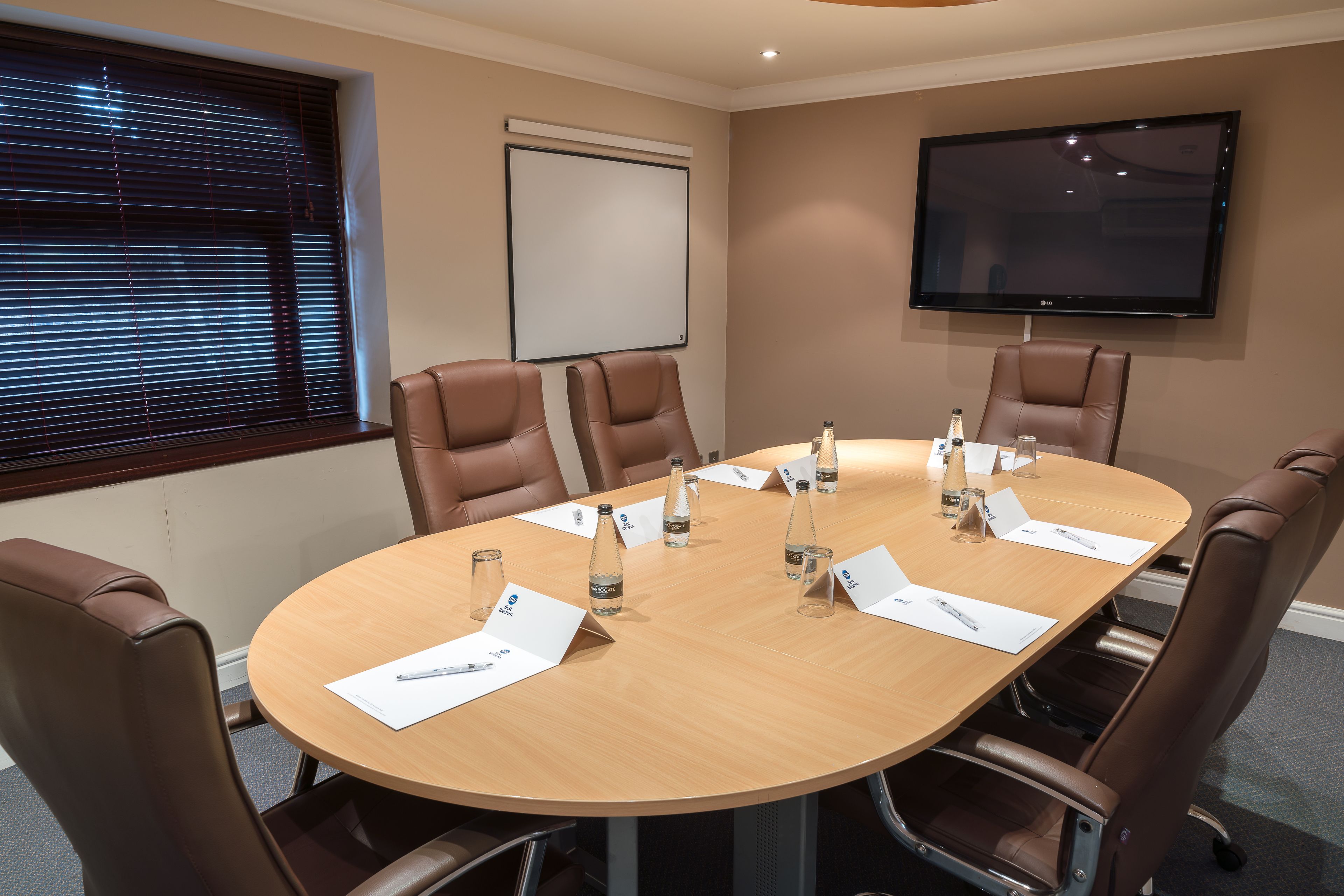 A conference room with a round table, brown leather chairs, a projector screen, and a flat-screen TV on the wall.