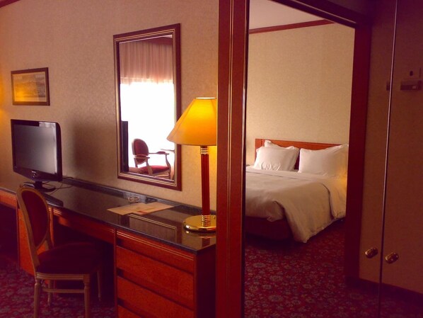 Quarto de hotel com escrivaninha, cadeira, televisão, luminária e cama com roupa de cama branca.