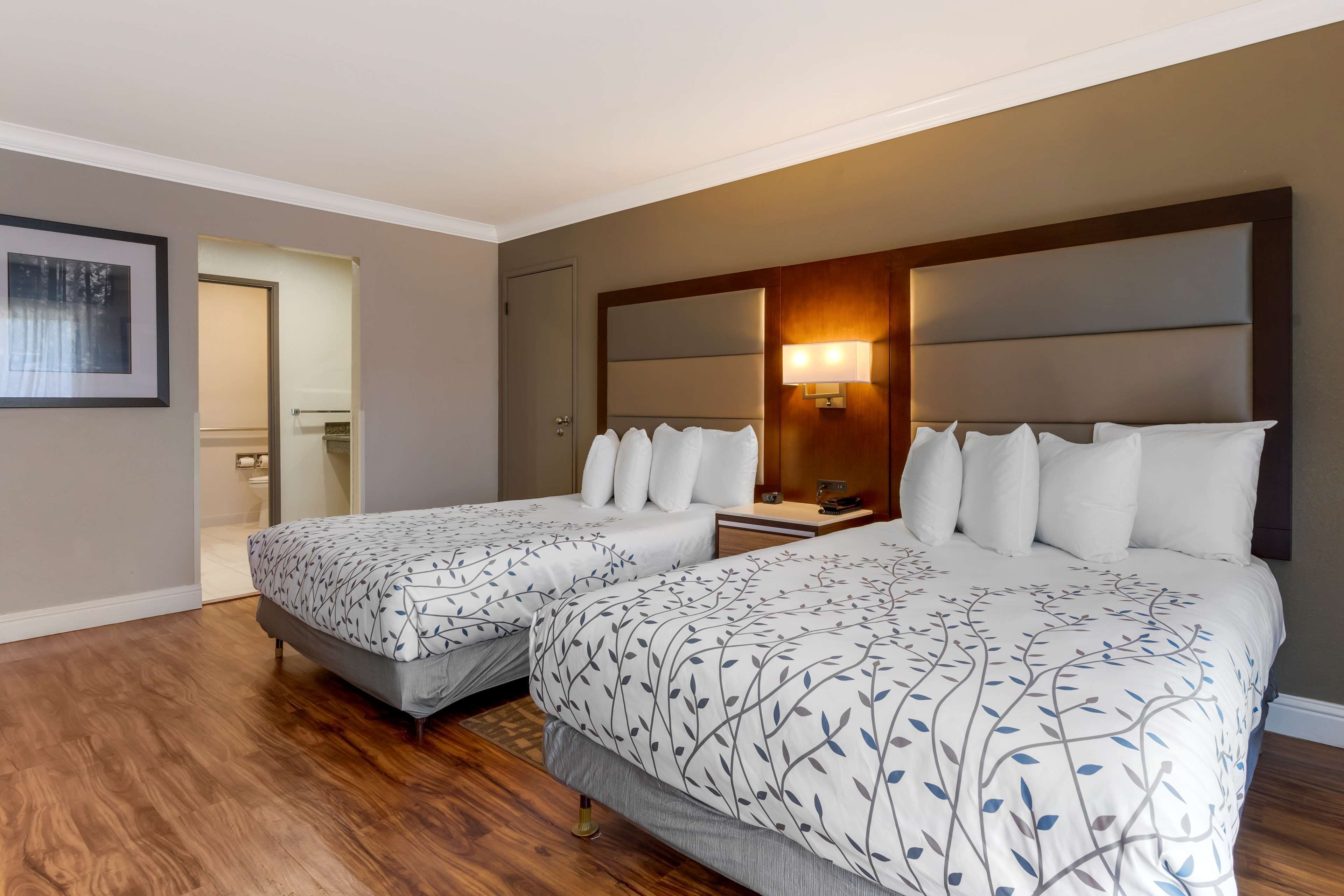 A hotel room with two beds, wooden flooring, a large mirror, and a nightstand with a lamp.