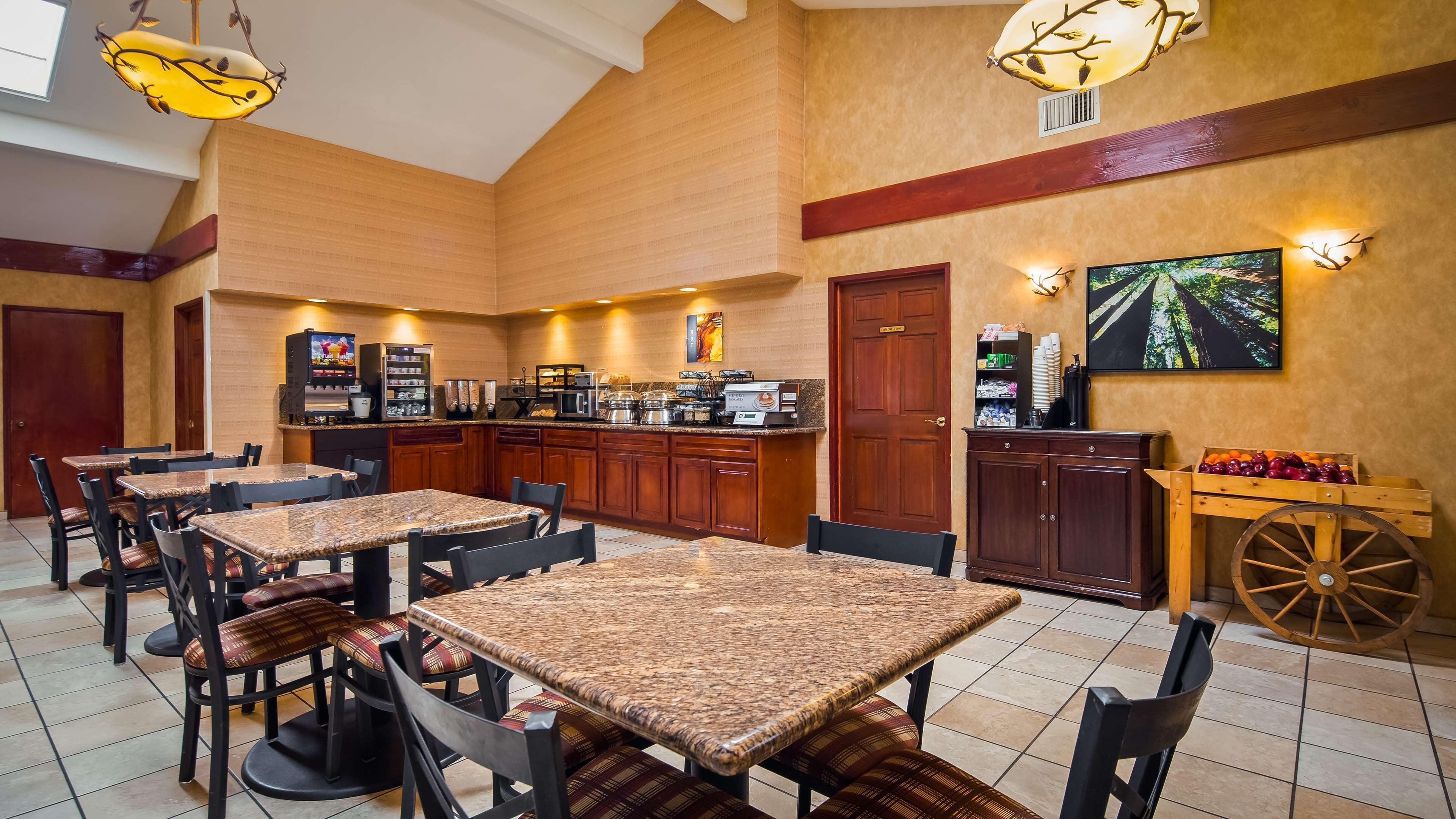 A restaurant with high ceilings, wooden paneling, and a central island with kitchen equipment. Tables and chairs are arranged around the island.