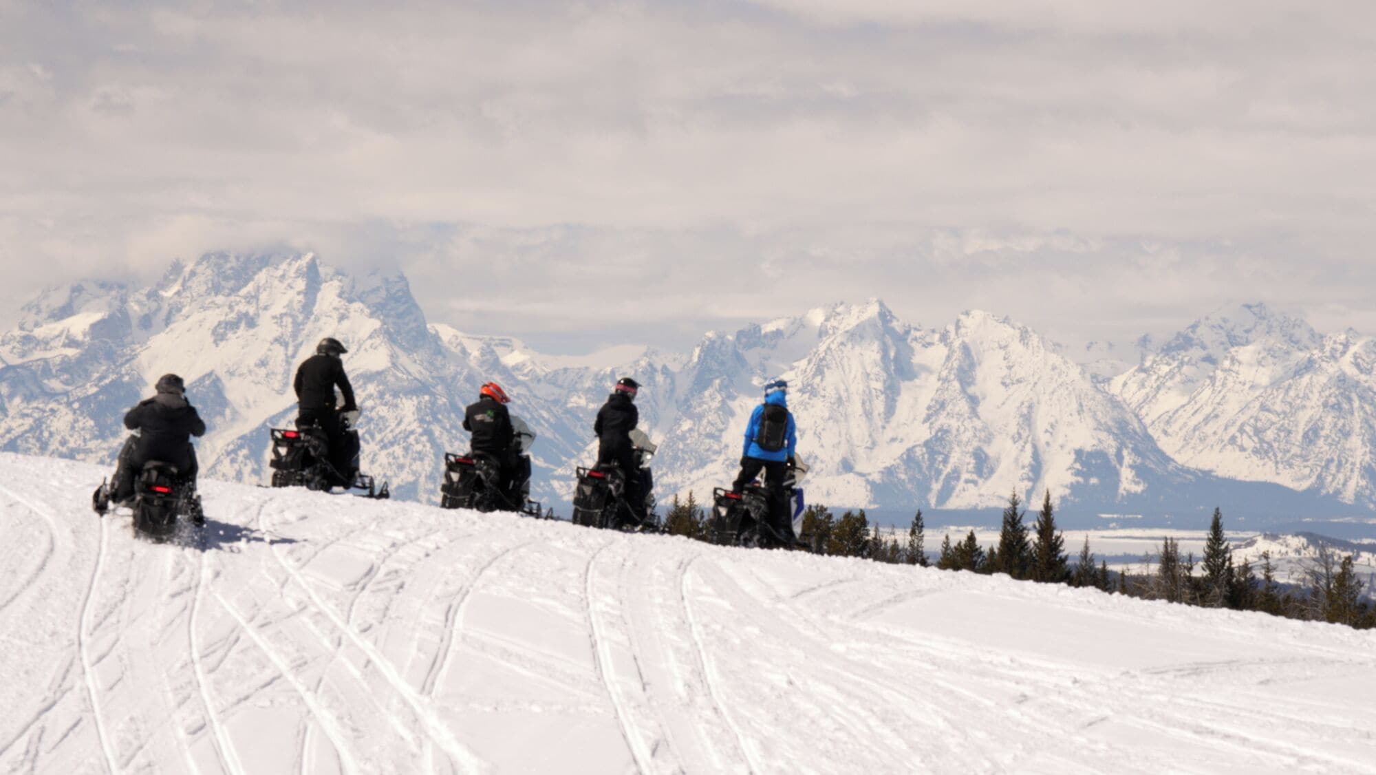 Een groep mensen op sneeuwscooters op een besneeuwde helling, met een bergketen op de achtergrond.