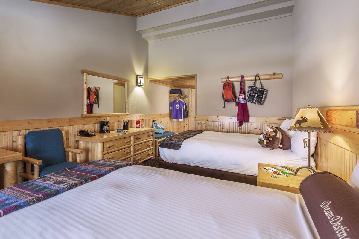 A cozy hotel room with two beds, a desk, and a chair. There are personal items and a mirror on the wall.