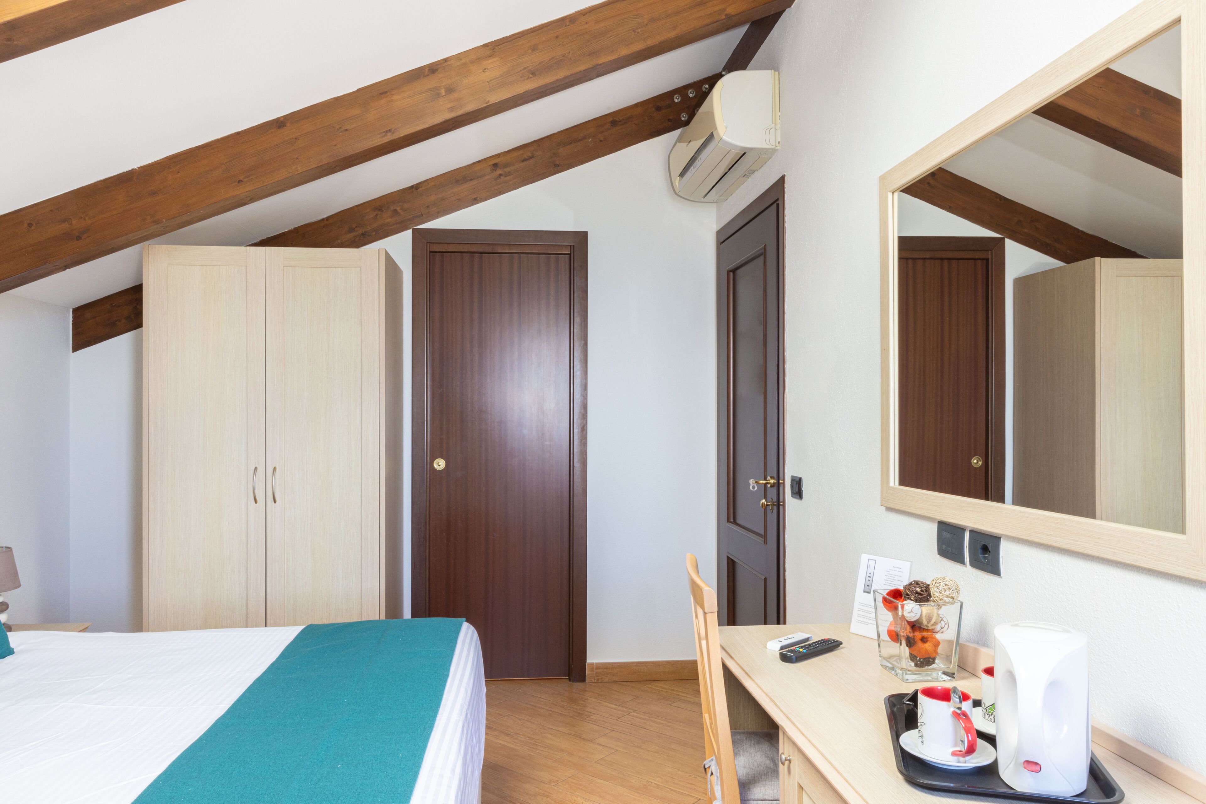 A bedroom with a bed, wooden wardrobe, a desk with a chair, a mirror, and an air conditioning unit.
