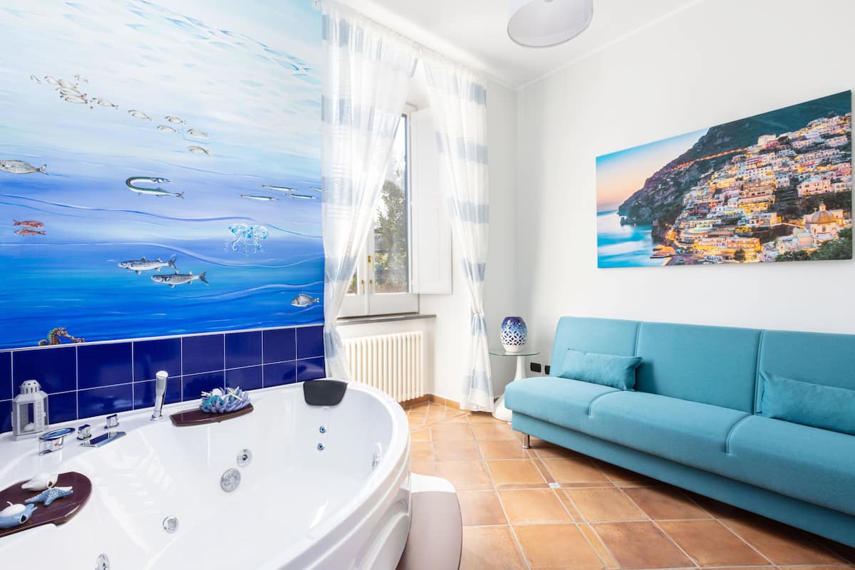 A bathroom with a large bathtub, a blue sofa, a painting of a coastal village, and a tiled wall with a seascape mural.
