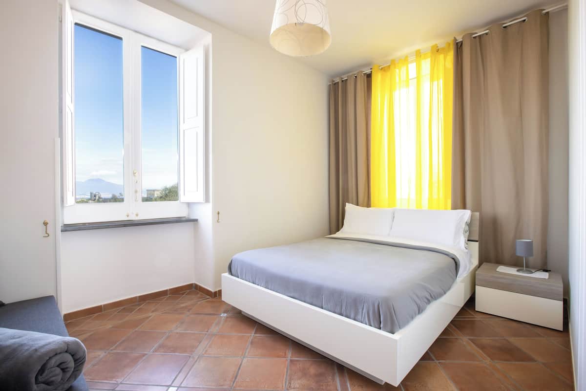 A bedroom with a bed, a nightstand, a lamp, a window with curtains, and a view of the outside.