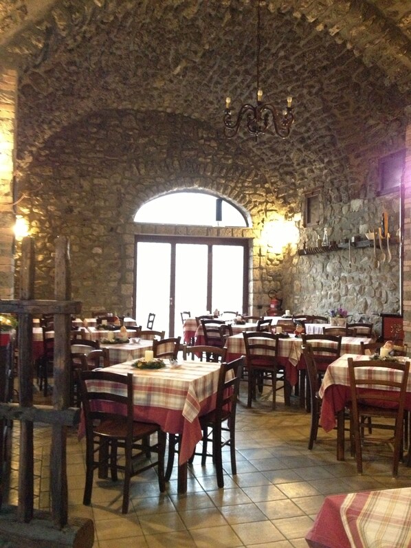Un restaurante con paredes de piedra y ventanas arqueadas, mesas dispuestas con manteles rojo y blanco, y sillas de madera.
