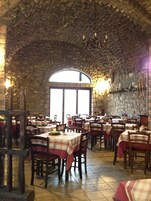 A restaurant with stone walls and arched windows, tables set with red and white tablecloths, and wooden chairs.