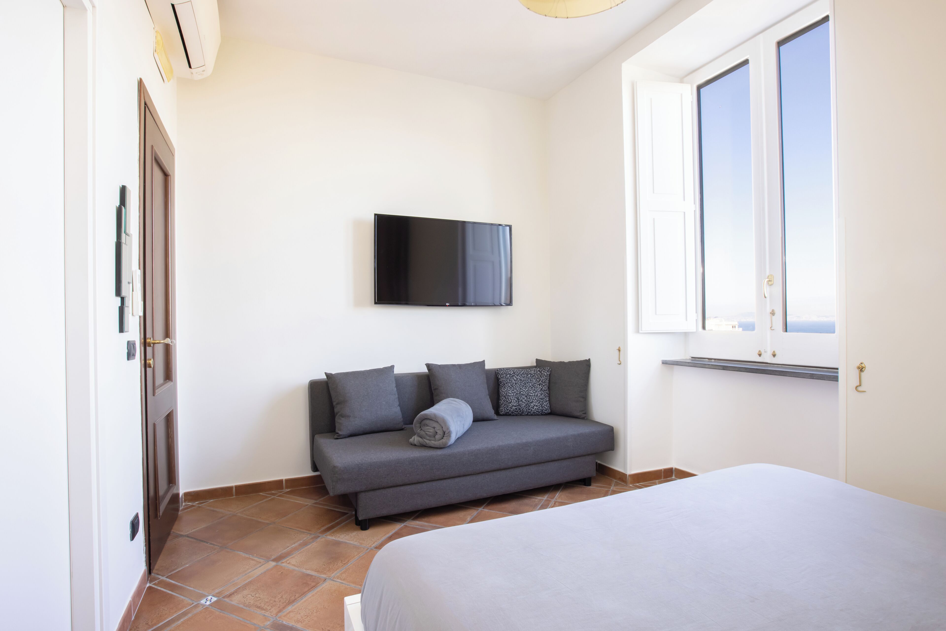 Un dormitorio con cama, sofá gris, televisor de pantalla plana y ventana.