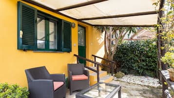 A covered outdoor seating area with wicker furniture and a glass table, adjacent to a yellow building with green shutters.