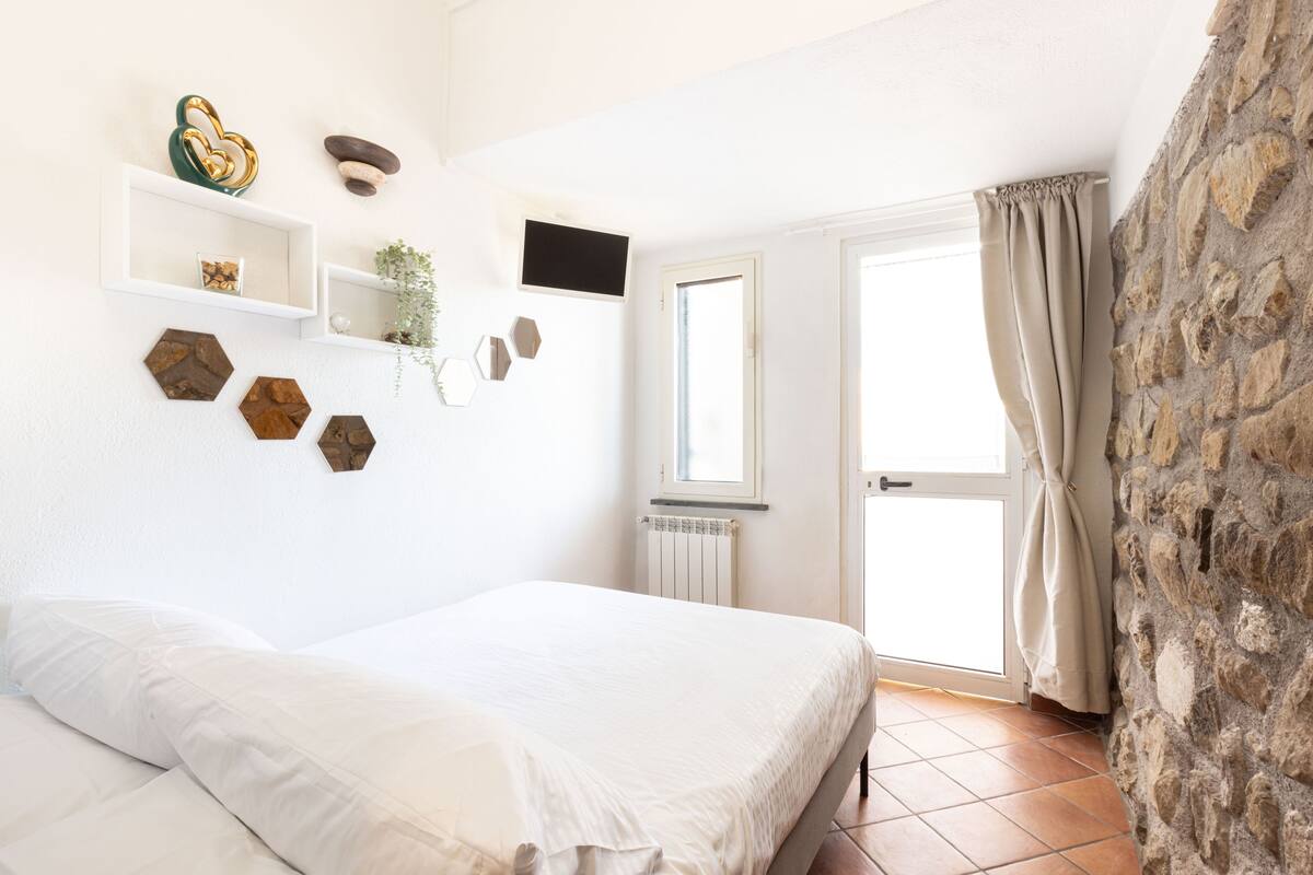 A bedroom with a bed, white walls, a stone wall, a window, and a door.
