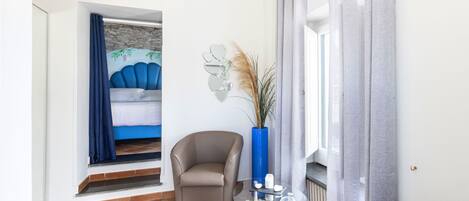 A modern living room with a glass coffee table, a comfortable armchair, and a view of a bedroom with a bed and a stone wall.