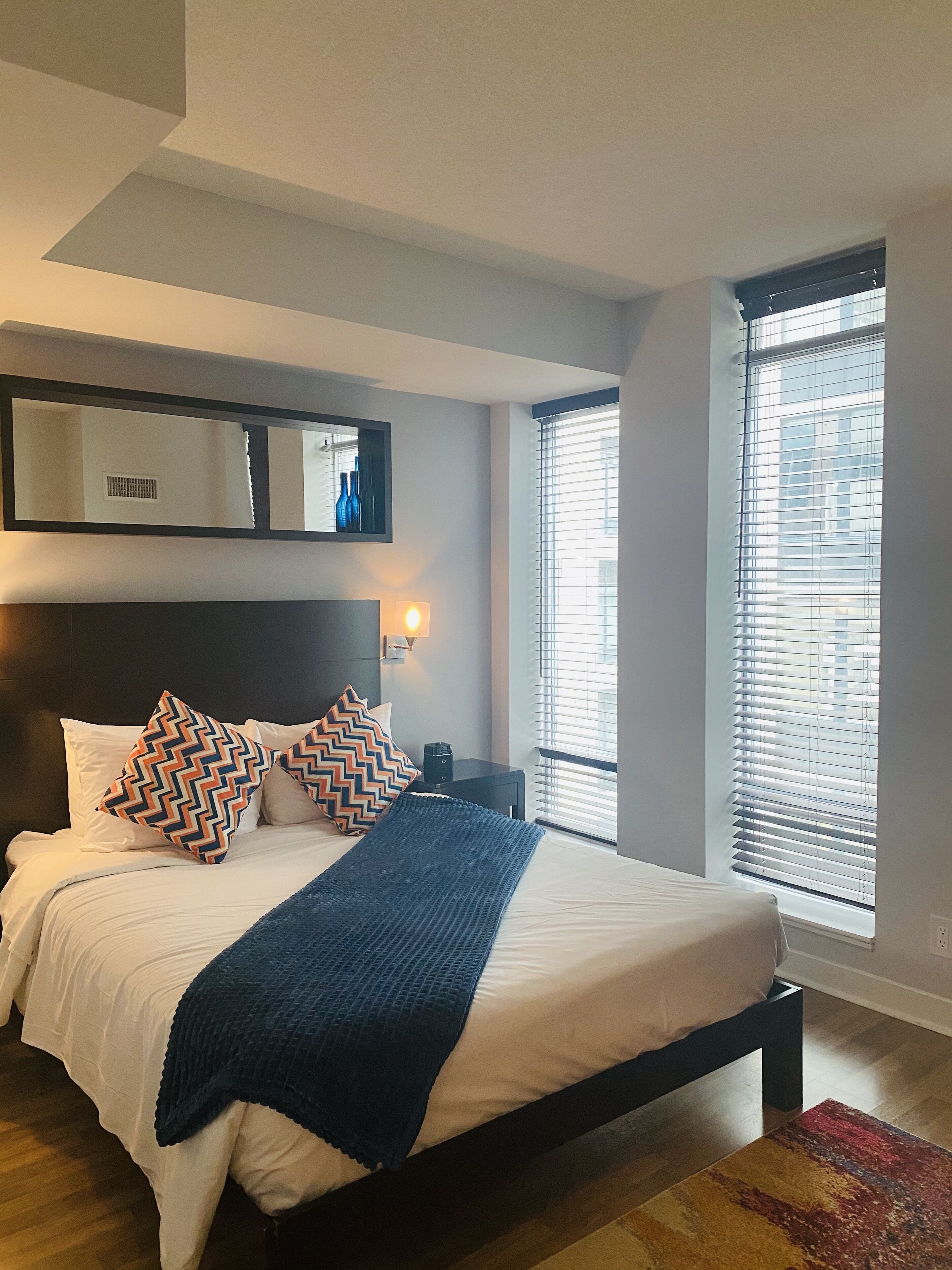 Een moderne slaapkamer met een groot bed, een donker hoofdbord, een spiegel aan de muur en een raam met jaloezieën.