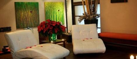 A room with two white cushioned chairs, a small table with a vase of red flowers, and a large window with bamboo decorations.
