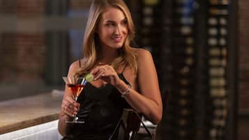A woman holding a martini glass with a lime wedge, seated at a bar with a brick wall background.