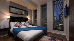 Une chambre d’hôtel moderne dotée d’un grand lit, offrant une vue sur la ville grâce à de grandes fenêtres du sol au plafond, d’un téléviseur à écran plat fixé au mur et d’une petite table de chevet avec une lampe.