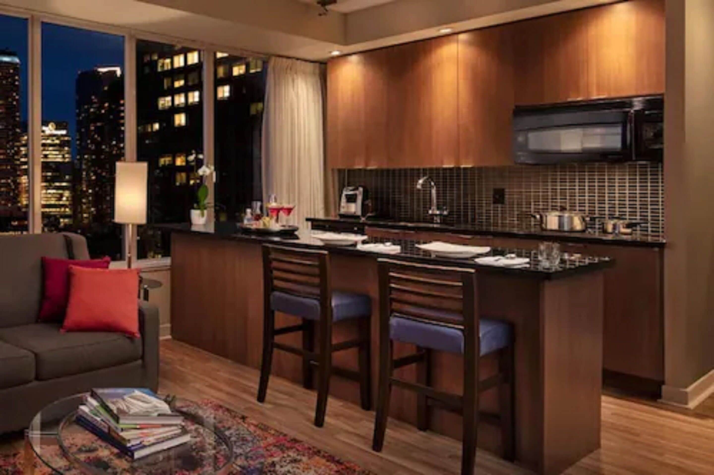 A modern kitchen with a bar area, large windows with a city view, and a cozy seating area with a sofa and a coffee table.