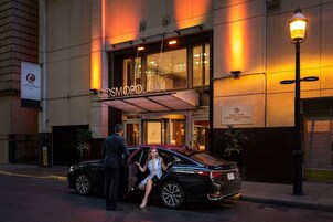 Entrada de hotel con un letrero que dice 'Cosmopolitan' y un hombre y una mujer saliendo de un coche negro.