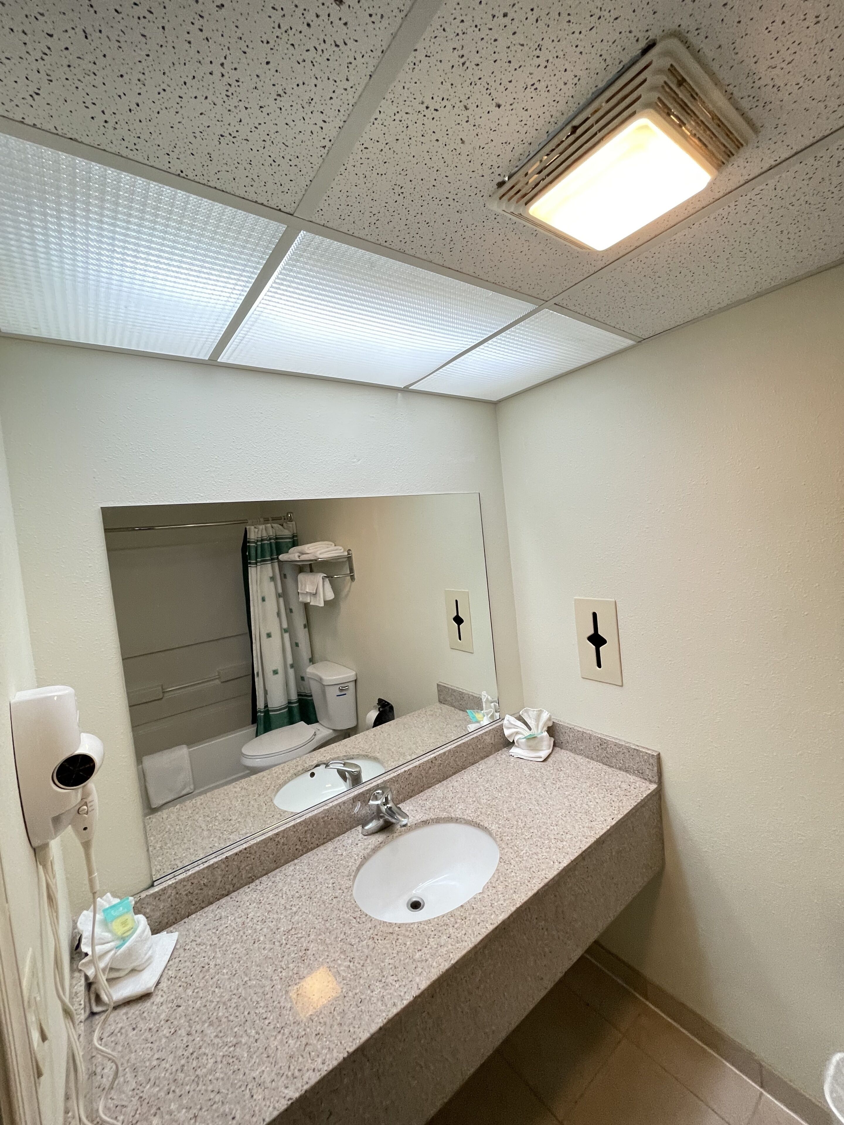 A bathroom with a large mirror, two sinks, a towel dispenser, and a toilet visible in the reflection.