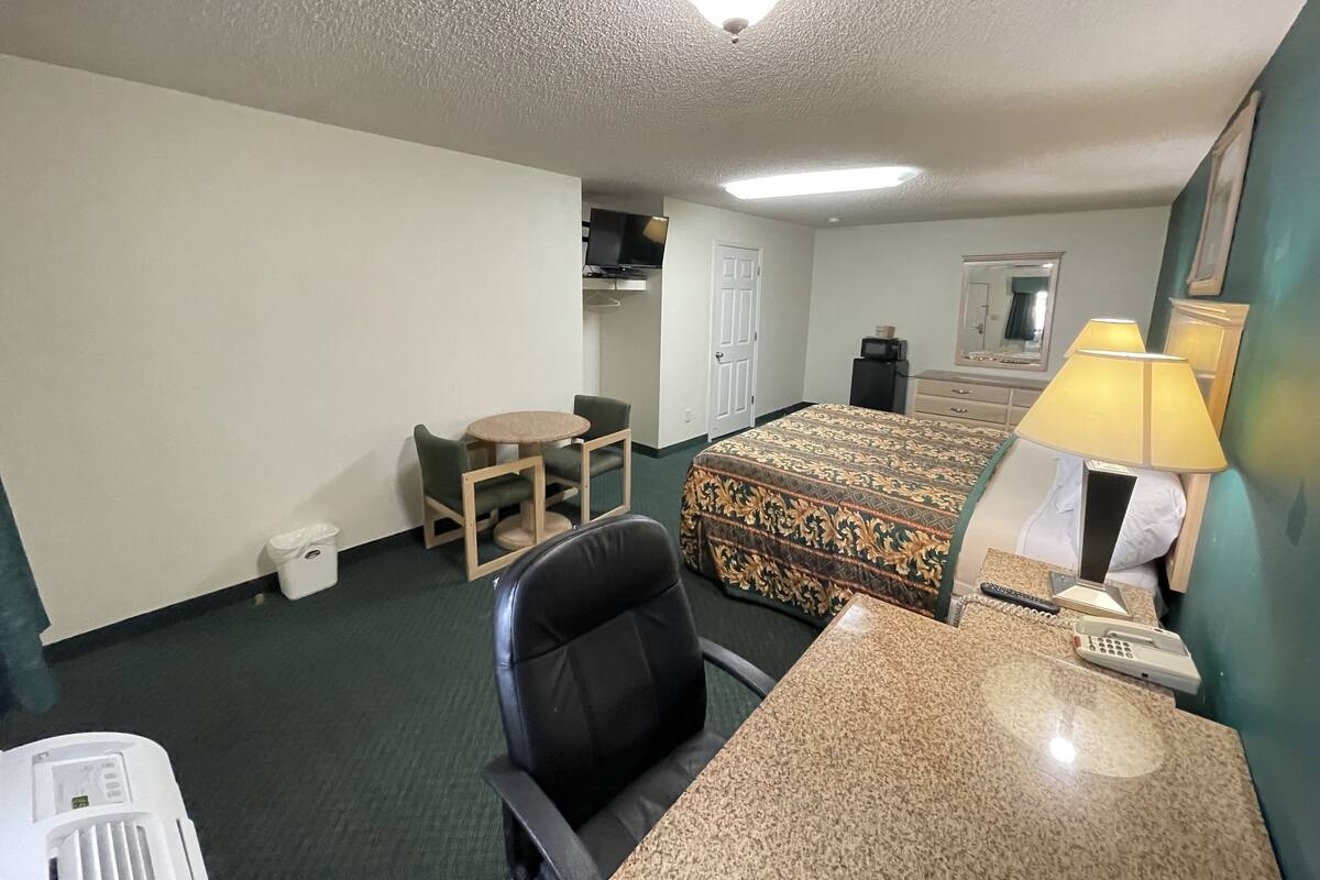 A hotel room with a bed, a desk with a lamp, a chair, a small table, a microwave, a dresser, and a mirror.