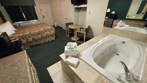 A hotel room with a large bed, a sitting area with a chair and table, a bathroom with a bathtub and sink, and a flat-screen TV mounted on the wall.