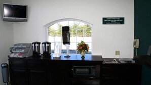 A hotel reception area with a flat-screen TV, a complimentary continental breakfast sign, a coffee machine, a microwave, and a vase with flowers.
