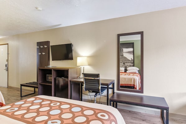Quarto de hotel com cama, escrivaninha com cadeira, televisão, luminária e um espelho grande.