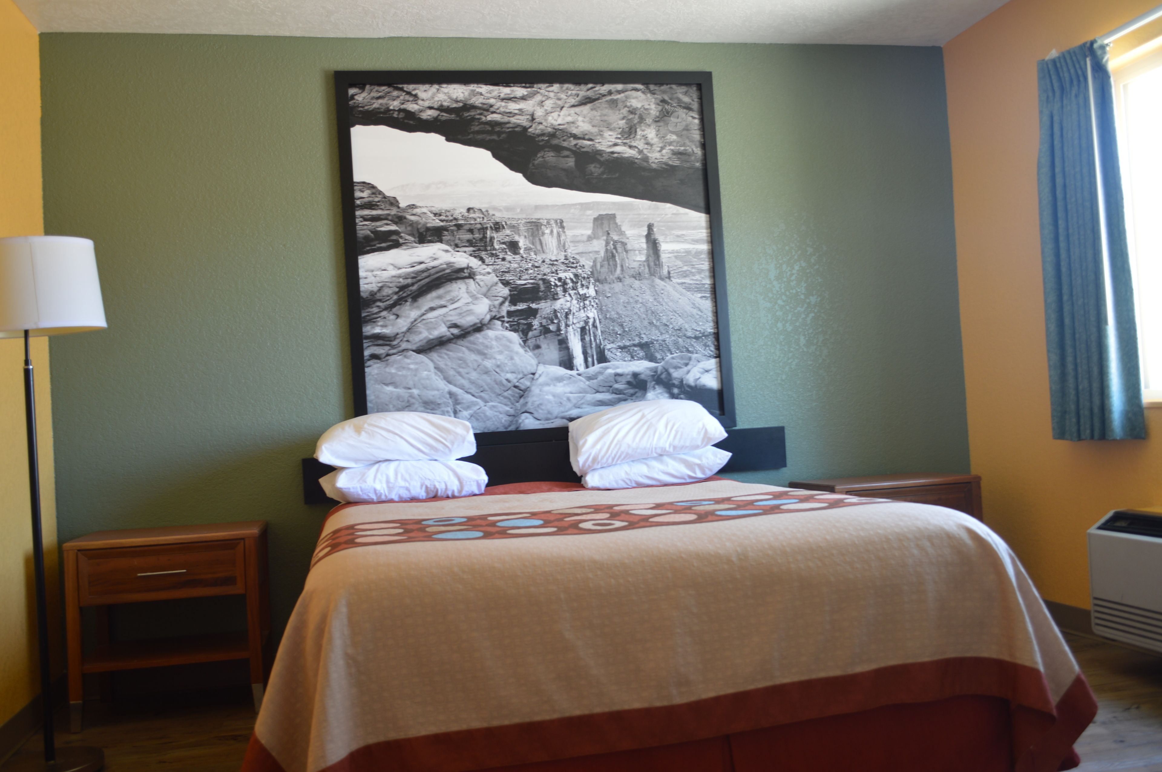A hotel room with a bed, a nightstand, a lamp, and a large framed picture on the wall.