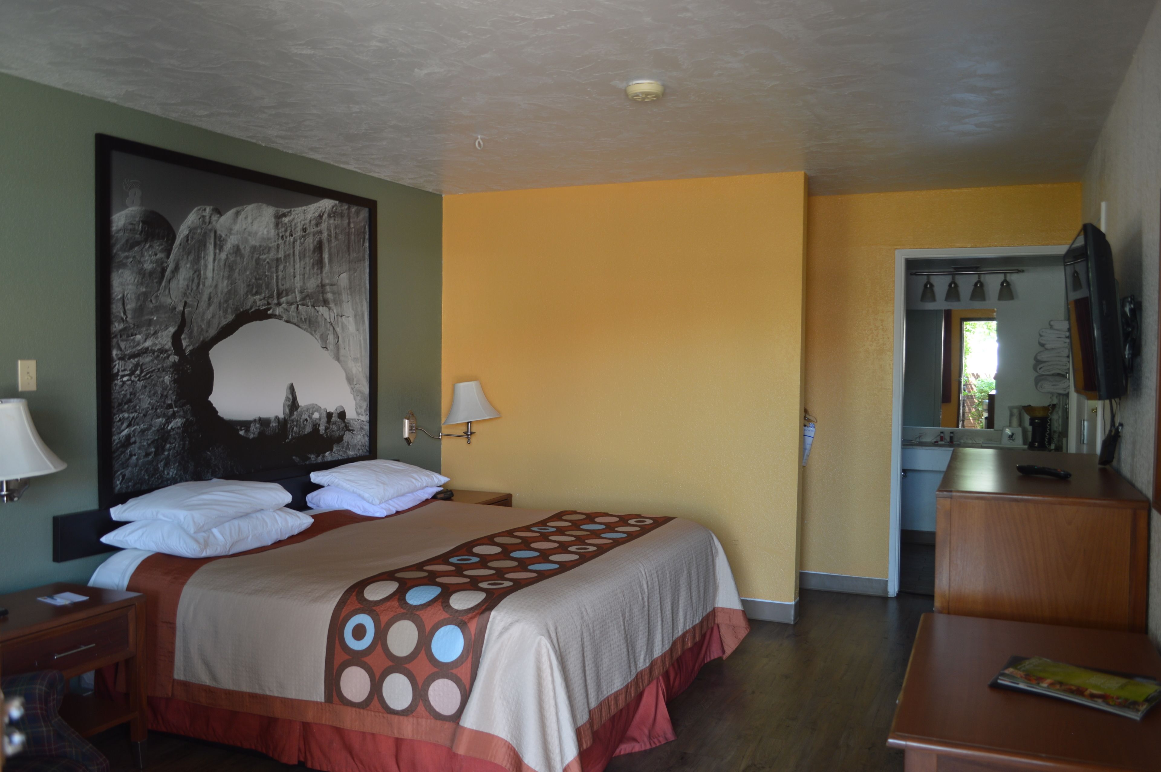 A hotel room with a bed, a desk, a chair, a bathroom visible through an open door, and a wall-mounted lamp.