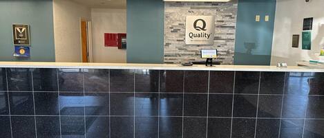 A hotel reception area with a marble counter, a 'Quality by Choice Hotels' sign, a digital welcome screen, and a plaque on the wall.