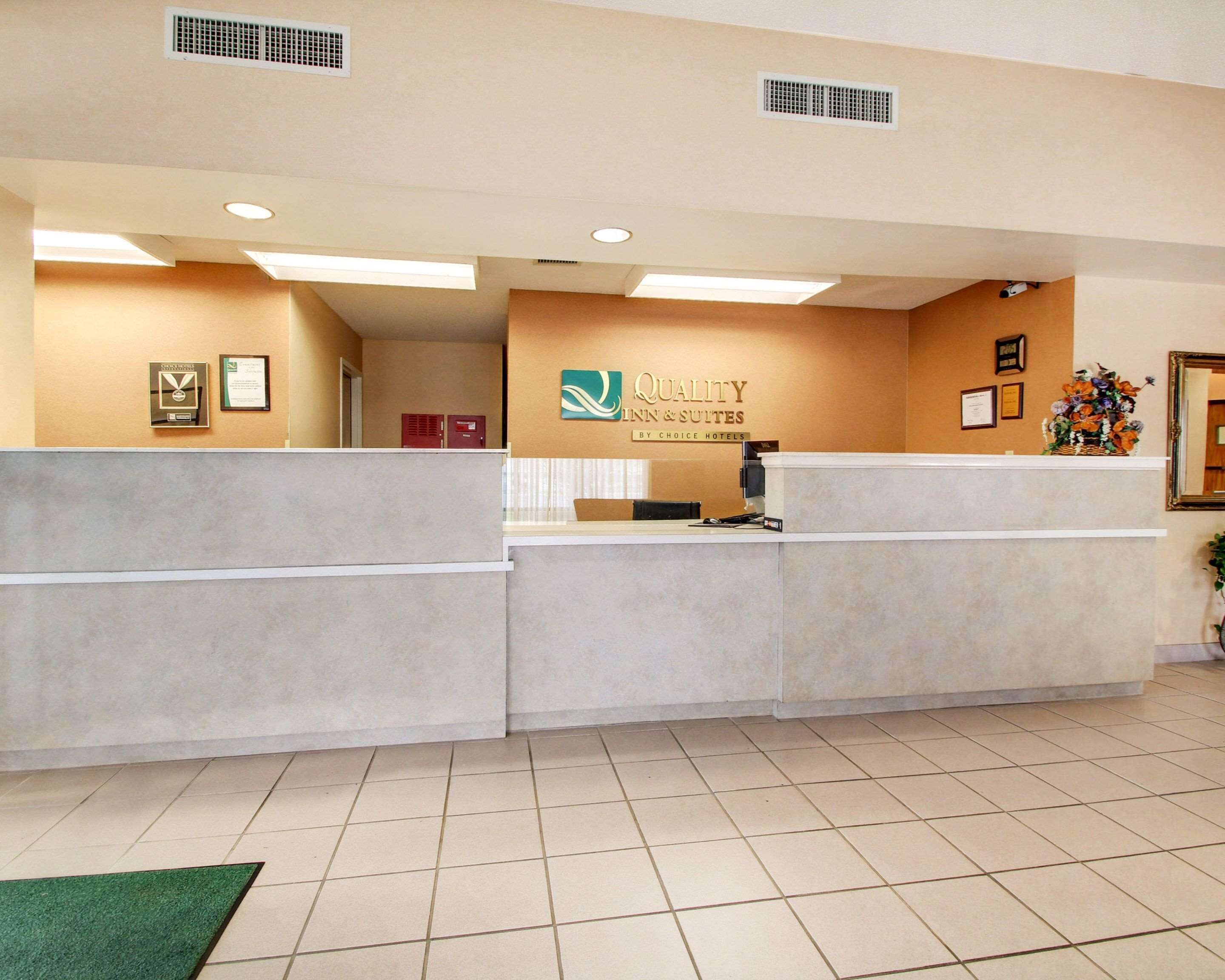 A hotel reception area with a reception desk, a wall-mounted sign for Quality Inn & Suites, and a framed certificate on the wall.