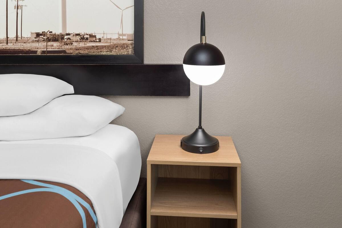 Ein ordentlich bezogenes Bett mit weißen Laken und einem braunen Kopfteil, eine schwarze Schreibtischlampe auf einem hölzernen Nachttisch und ein gerahmtes Bild von Windkraftanlagen an der Wand.