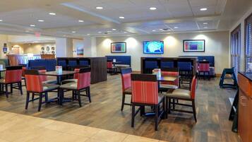 A hotel dining area with multiple tables and chairs, a reception desk, and a television screen.