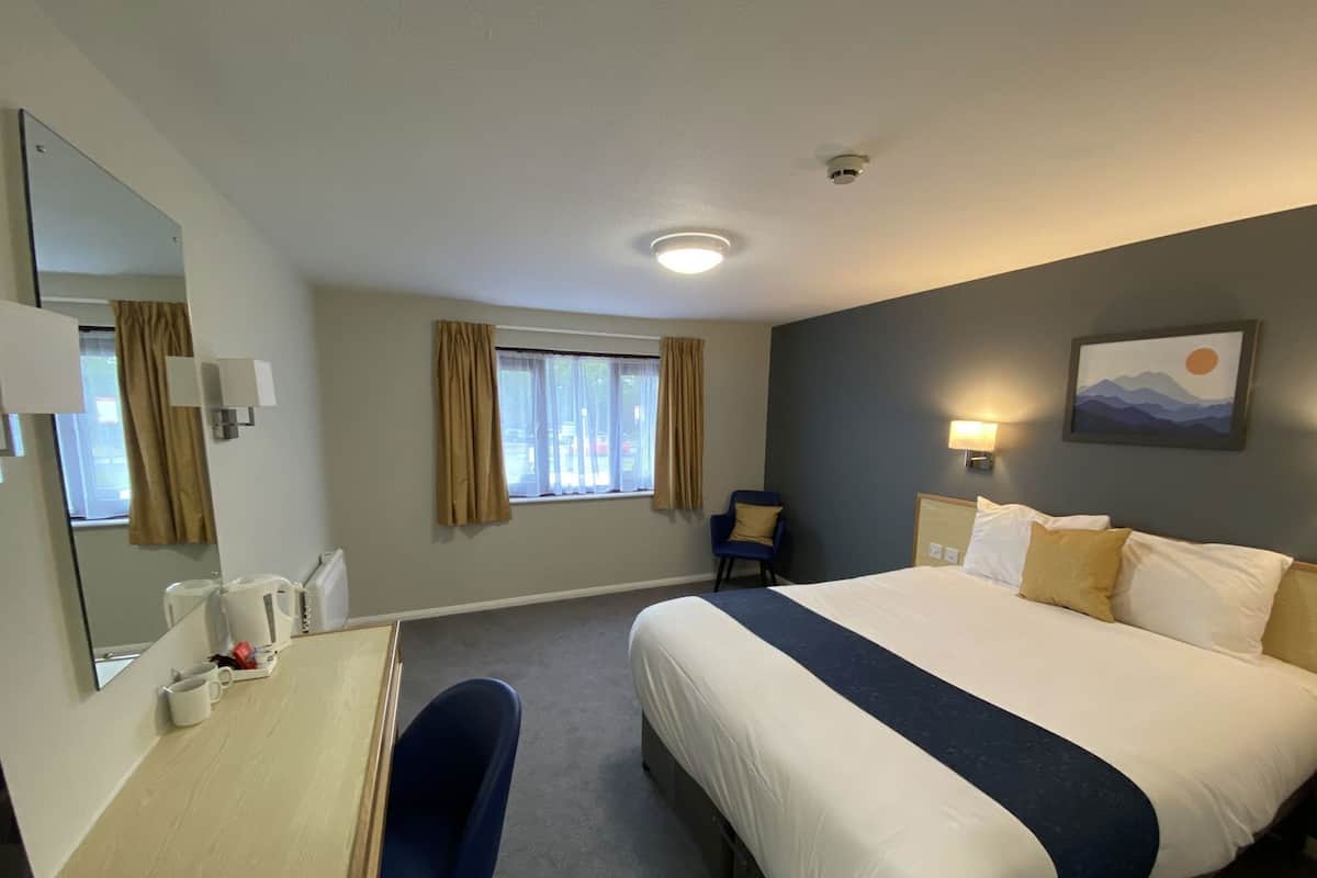 Ein Hotelzimmer mit Bett, Schreibtisch, Stuhl, einem Fenster mit Vorhängen, einer Wandlampe und einem Gemälde.