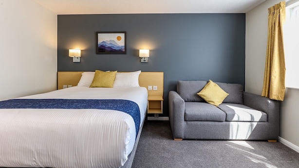 Een hotelkamer met een groot bed, een grijze bank, twee wandlampen en een schilderij van bergen.