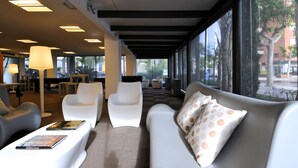 A modern hotel lobby with white sofas, a coffee table, and a large window offering a view of the street.