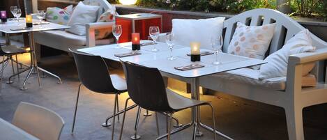 Un espacio al aire libre para comer con mesas, sillas, velas y un banco con almohadones.