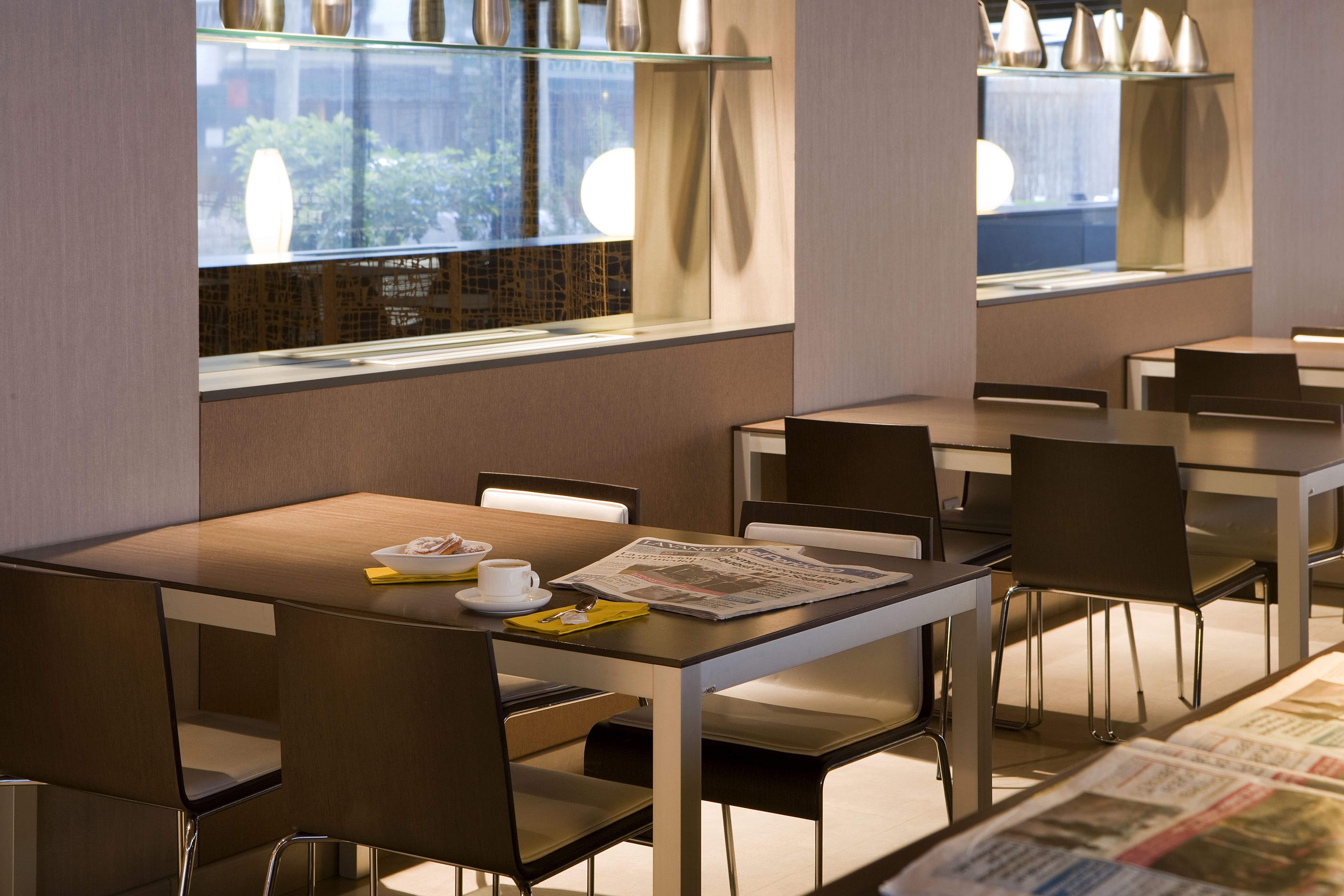 A modern dining area with tables and chairs, newspapers on the tables, and a window with a view of greenery.