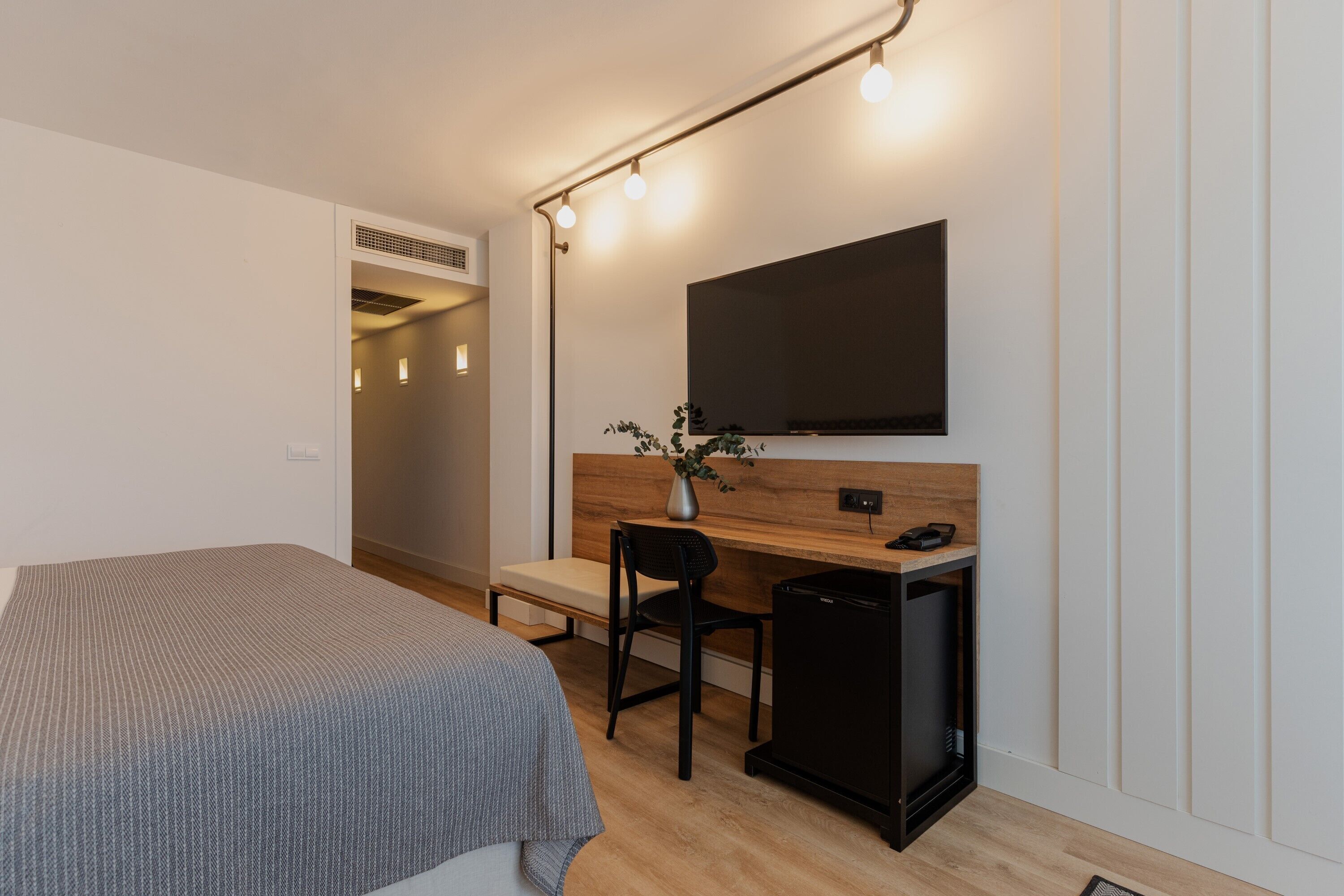 Una habitación de hotel moderna con una cama, un escritorio, una silla, un pequeño mueble negro, un televisor de pantalla plana montado en un panel de madera y una planta sobre el escritorio.