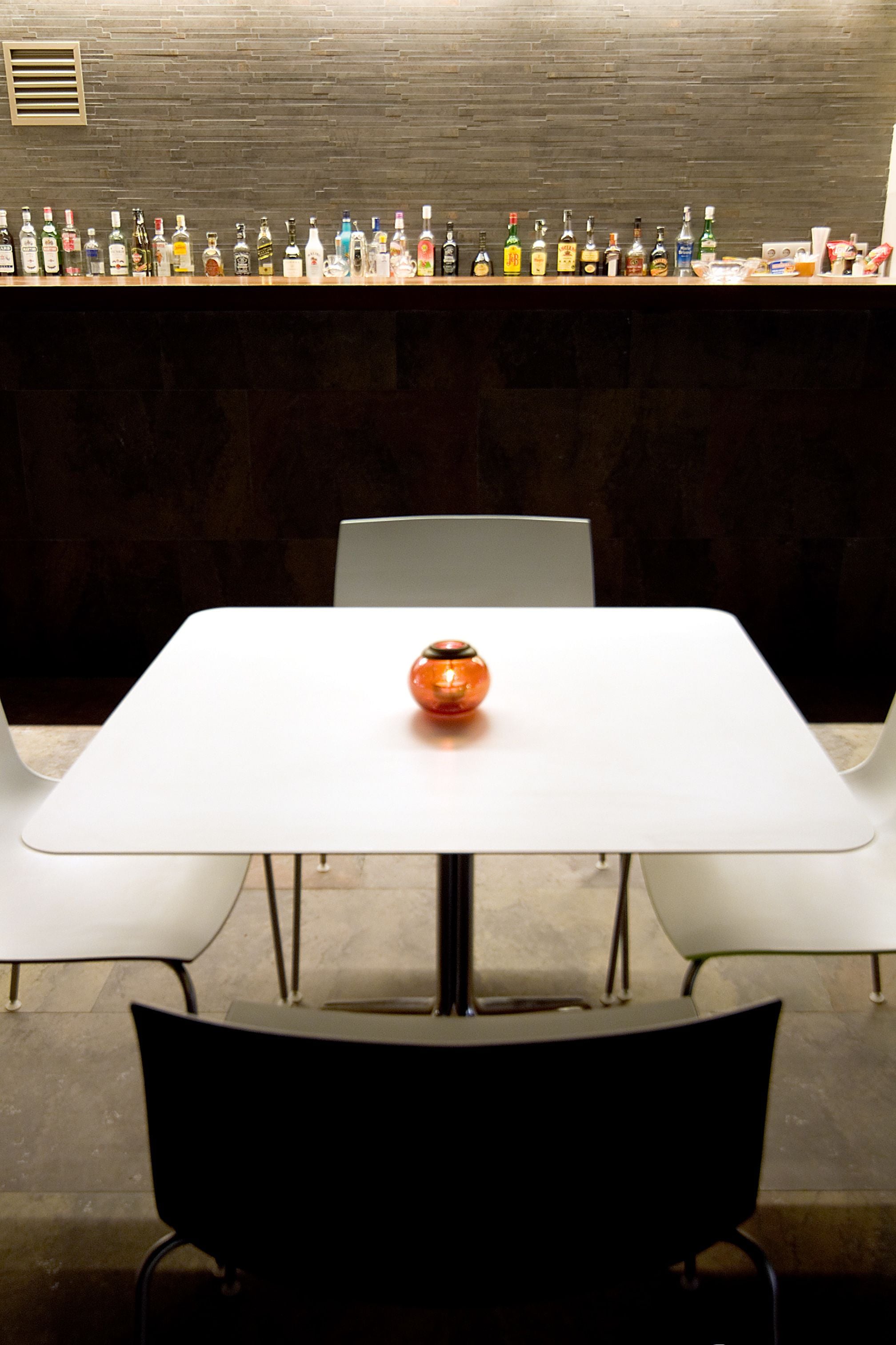 Una zona de bar con una larga hilera de botellas de licor en la pared y una mesa blanca con un solo jarrón naranja en el centro.